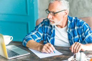portrait senior man writing notes using laptop table